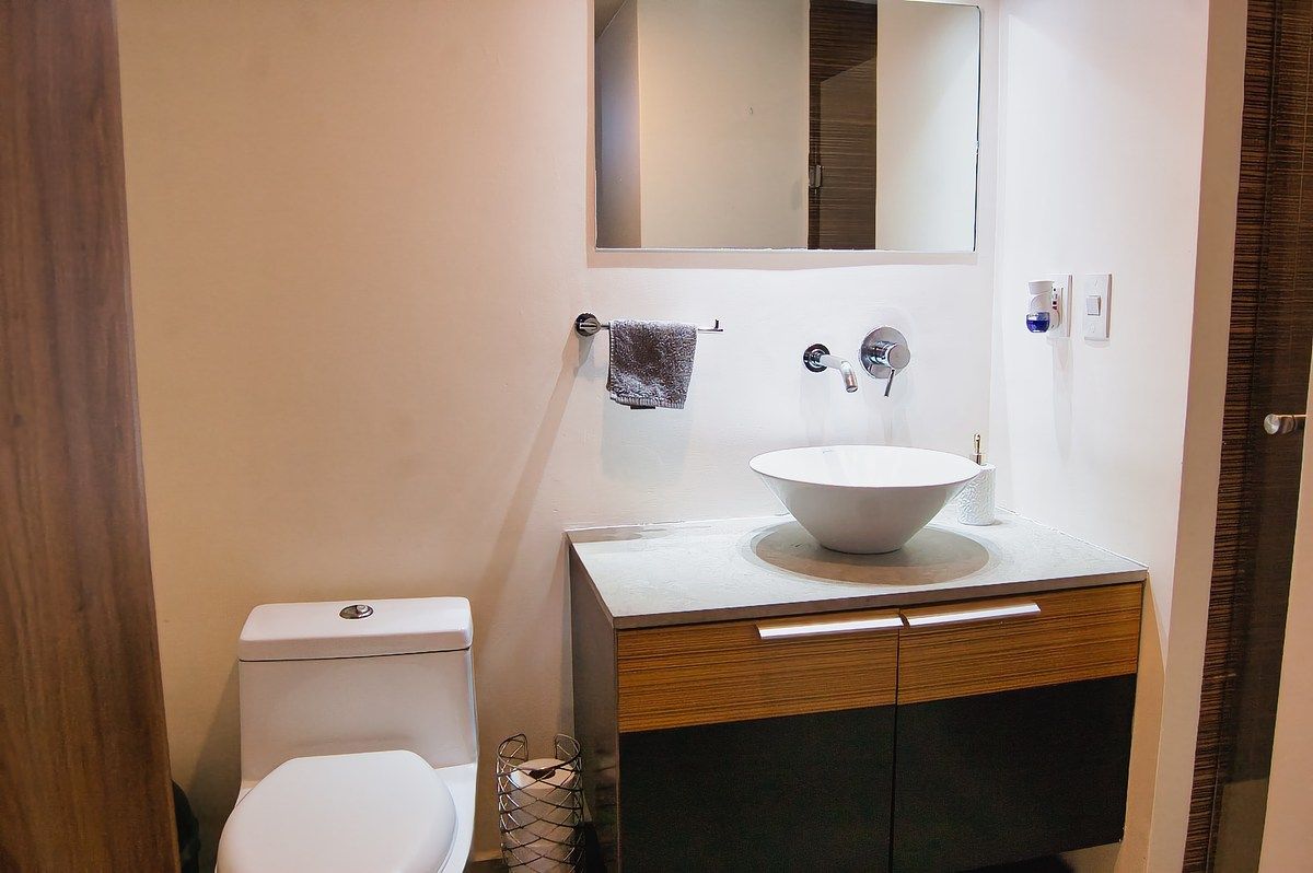 Baño principal con lavabo tipo ovalín cerámico sobre encimera gris, mueble flotante de madera oscura y wc blanco en espacio limpio.