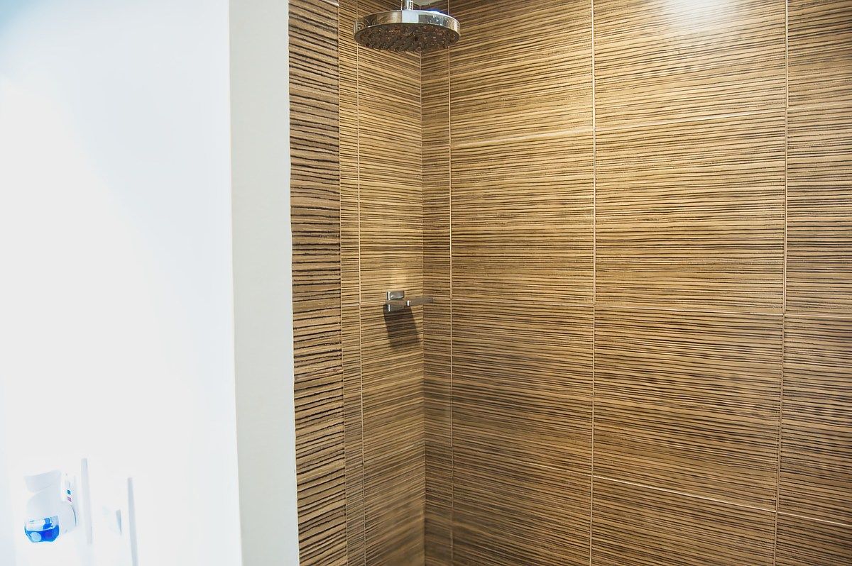 Detalle de la zona de ducha con regadera de lluvia de acero inoxidable y paredes revestidas con azulejo texturizado tipo madera horizontal.