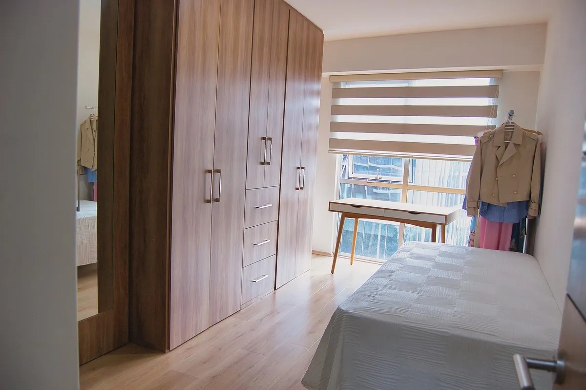 Vista de recámara secundaria iluminada con clóset empotrado de madera de piso a techo, piso laminado, escritorio pequeño junto a la ventana con persianas cebra y cama individual.