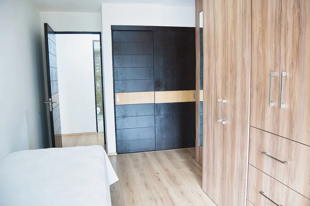 Vista interior de recámara secundaria con pisos de madera clara, clóset empotrado de madera con cajonera y puertas deslizables oscuras de diseño moderno.