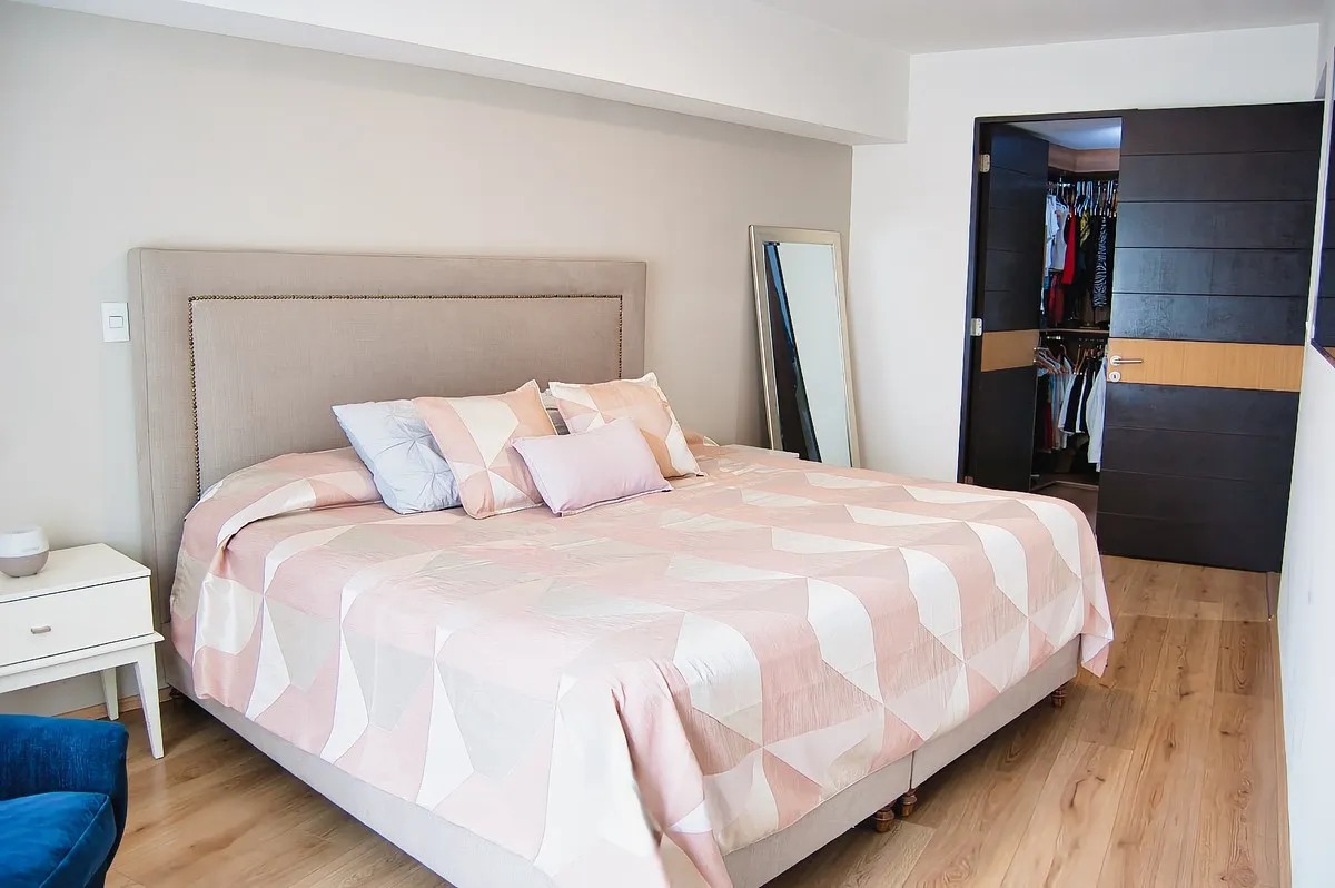 Recámara principal espaciosa con cama King Size, cabecera tapizada en color beige, buró blanco y gran puerta abierta de madera oscura que revela el walk-in closet.