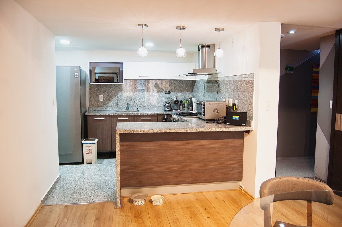Cocina integral abierta con barra de granito gris, gabinetes de madera oscura, electrodomésticos de acero inoxidable (refrigerador, estufa, campana) y lámparas colgantes modernas.