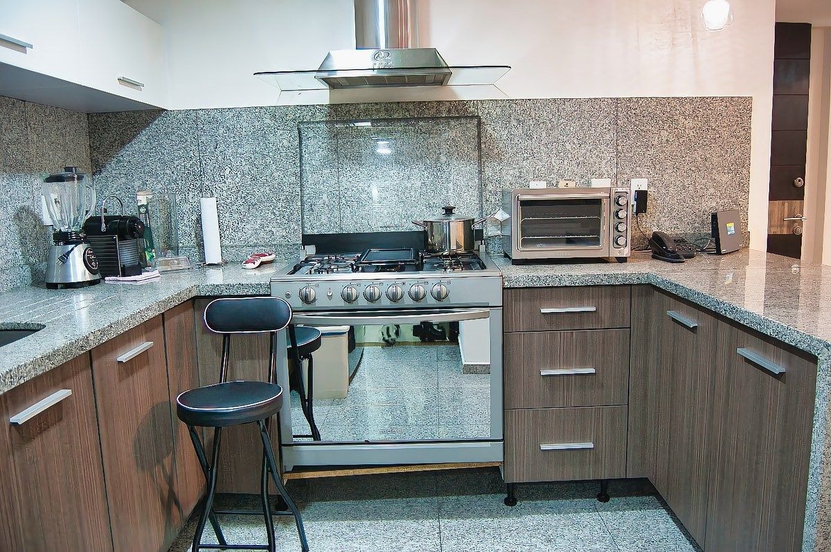 Zona de cocción con estufa de acero inoxidable de 5 quemadores, campana extractora de vidrio, horno tostador, gabinetes de madera oscura y granito gris.