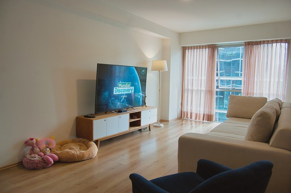 Sala de estar con piso laminado de madera, un gran mueble de TV blanco y madera, sofá claro, lámpara de pie moderna y ventanal de piso a techo con cortinas claras.
