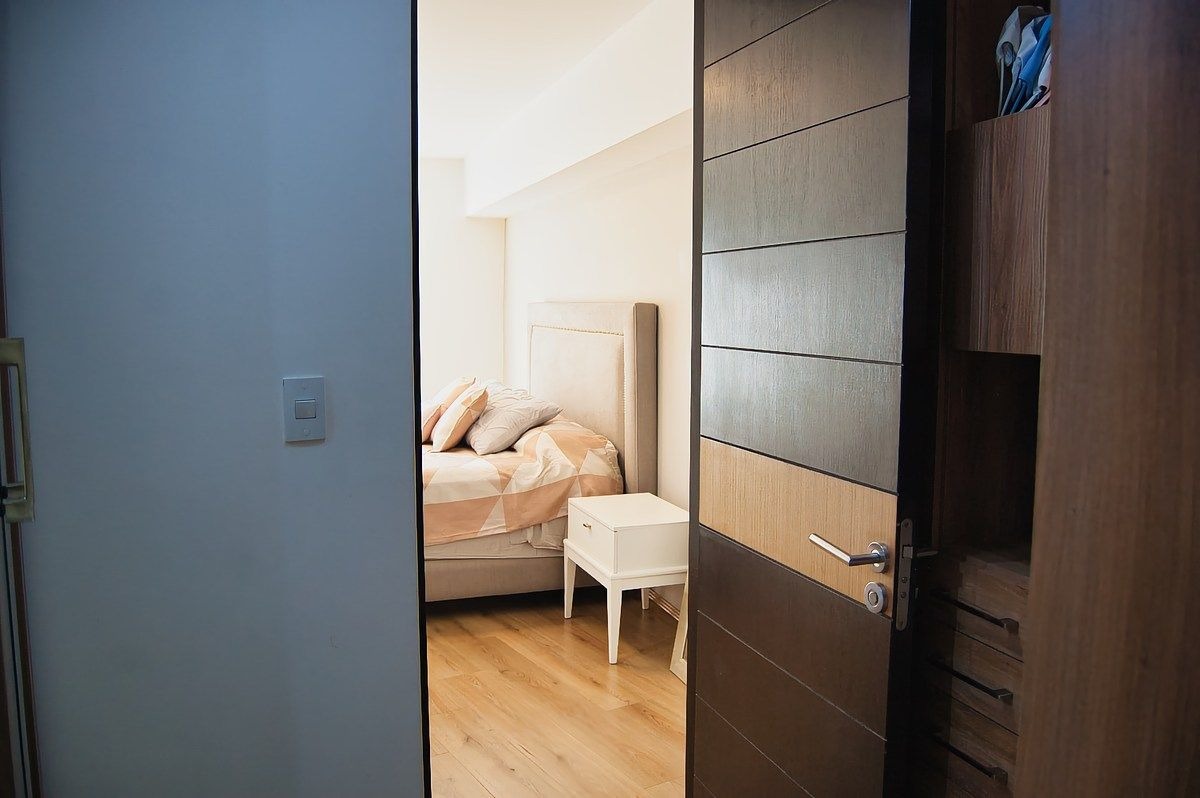 Vista de la recámara principal enmarcada por la puerta de madera oscura del vestidor, mostrando la cama con ropa clara, buró blanco y pisos de madera.