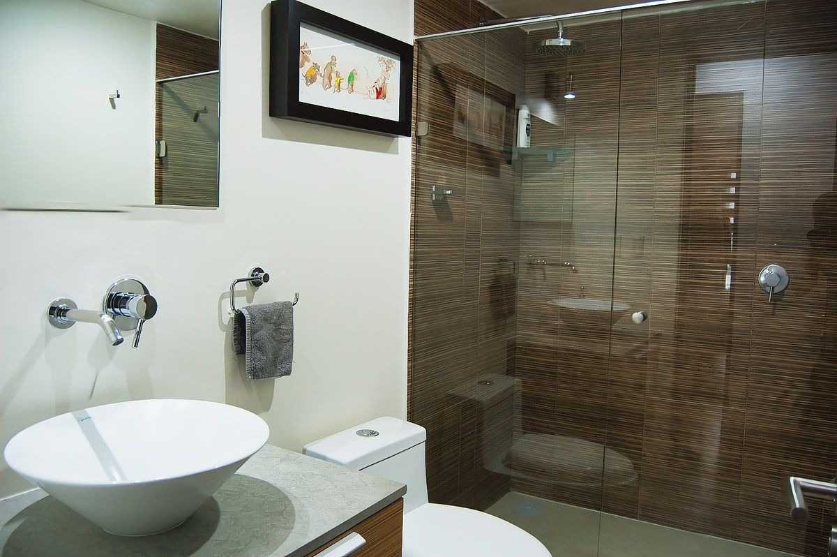 Baño secundario moderno con lavabo de ovalín sobre meseta de granito gris, cancel de vidrio templado en la regadera, azulejos tipo madera en la zona húmeda y regadera de lluvia.