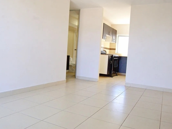 Vista panorámica de la estancia principal con pisos de cerámica blanca, mostrando el acceso a la cocina semi-equipada a la derecha y al baño secundario al fondo, en departamento residencial