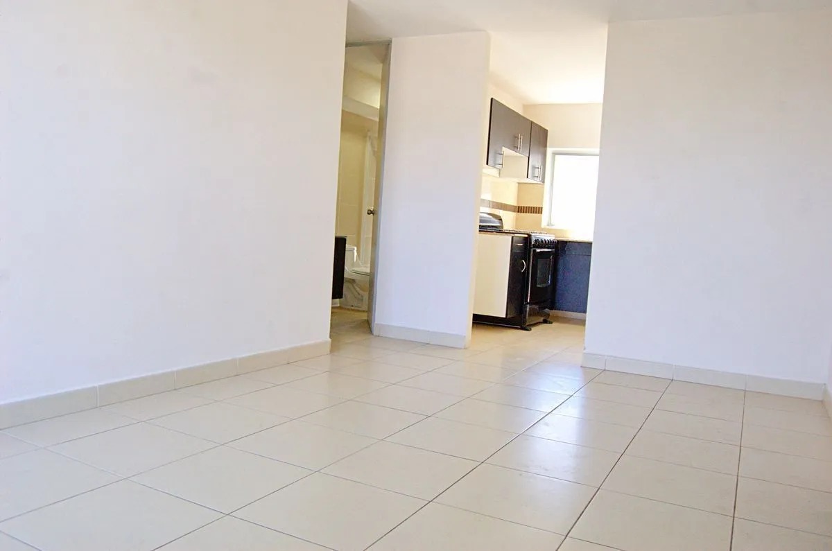 Vista panorámica de la estancia principal con pisos de cerámica blanca, mostrando el acceso a la cocina semi-equipada a la derecha y al baño secundario al fondo, en departamento residencial