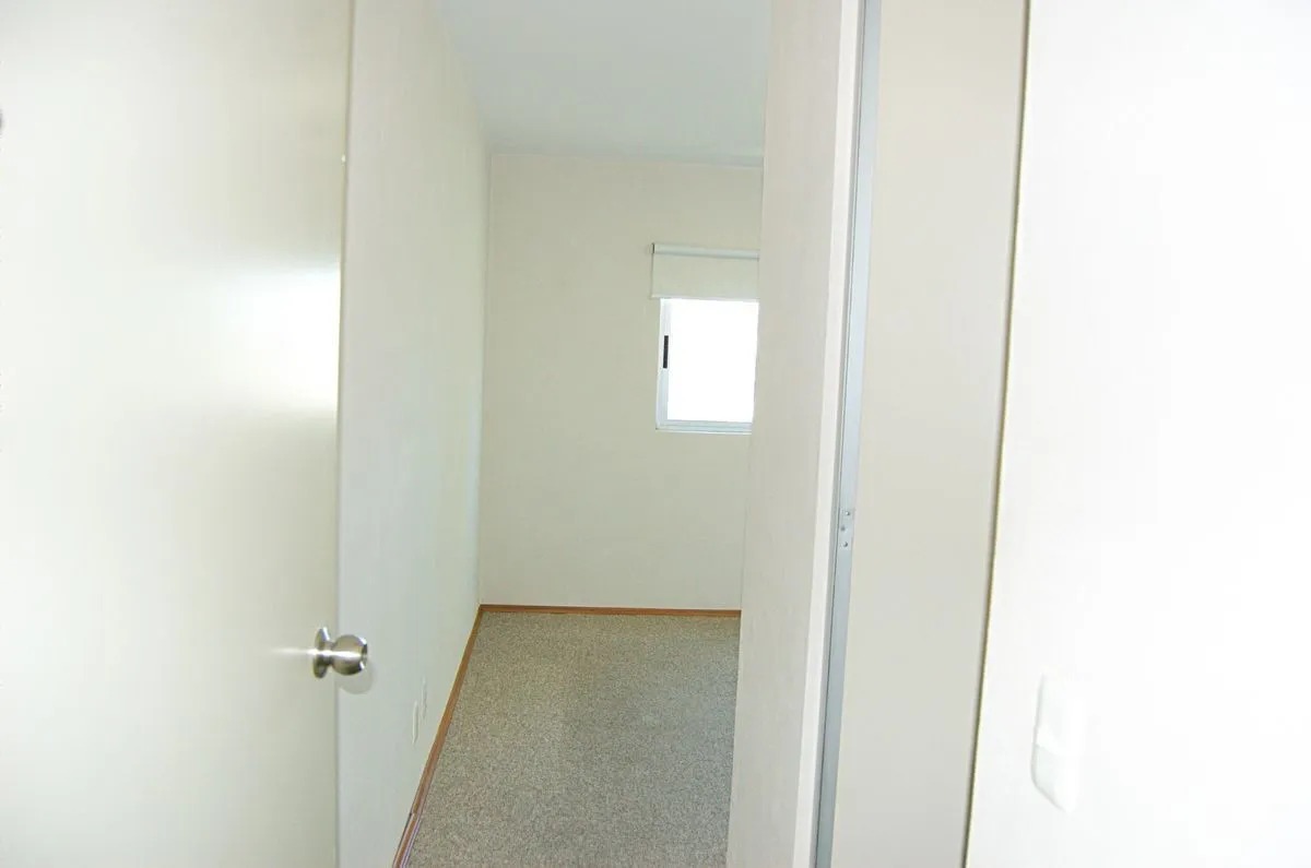 Vista a la recámara principal desde la puerta, mostrando el suelo con alfombra gris, ventana con persiana black out, y a la derecha la puerta para el baño